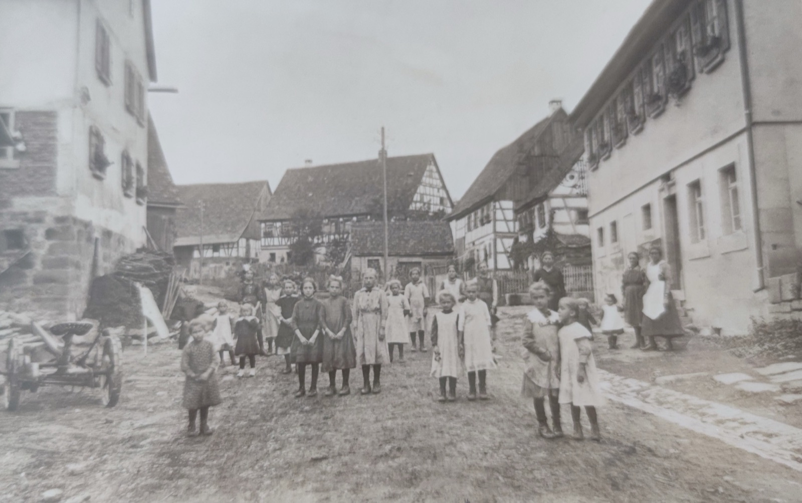 Altes Schwarz-Weiß-Foto: Kinder und Frauen stehen auf der unbefestigten Dorfstraße zwischen Fachwerkhäusern
