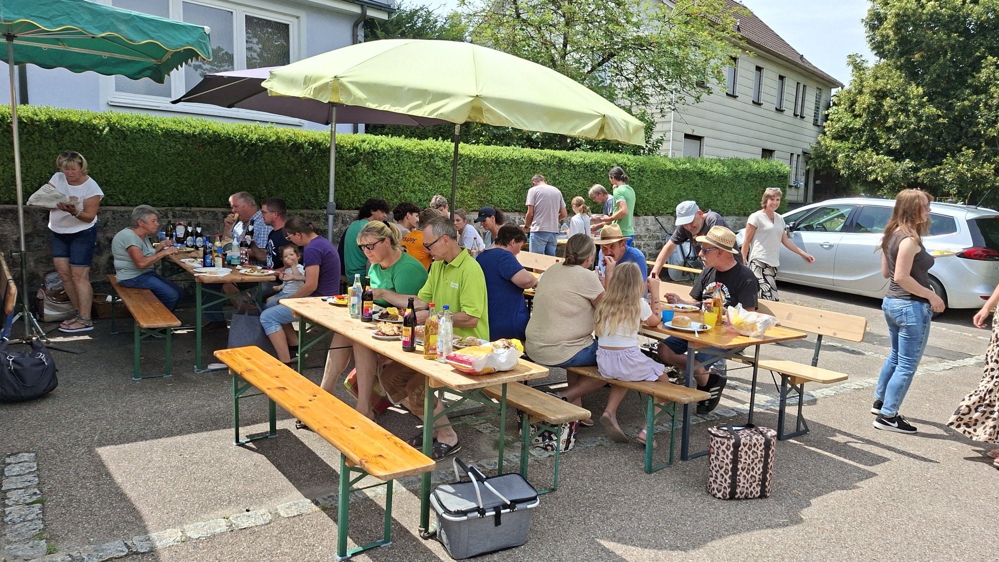 Gäste beim Essen an Bierbänken unter Sonnenschirmen vor einer Hecke