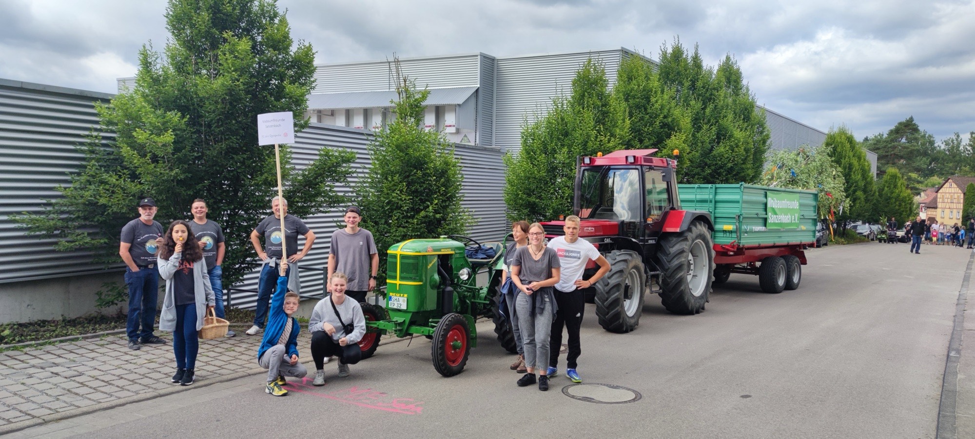 Gruppe der Maibaumfreunde mit Schild, grünem Deutz-Oldtimer und rotem Traktor mit Anhänger